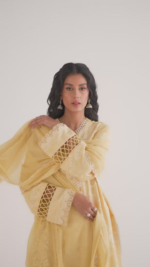 The Butter Yellow Set by Mannam is a soft lemon-hued Pima cotton three-panel kameez featuring ivory embroidery, a scalloped lace insert in the centre panel and loop fabric detailing on the sleeves. It comes with pin-tucked trousers with pleats and lace at the cuff, and a flowing georgette dupatta with embroidered borders and floral corner motifs.