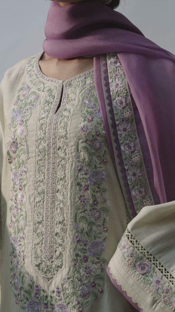 The Pistachio Set by Mannam is a pale pistachio green pure silk A-line long kameez with embellishments on the neckline, sleeves and hem, lace finishings and a distinctive purple border that peeks through the hem. It comes with pure silk scallop-edged trousers with lace finishings and a pure organza dupatta with an embroidered border.