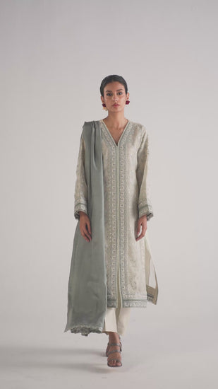 The Frost Set by Mannam is a pure organza kameez with an open-bottom silhouette, intricate embroidery and sequin embellishments throughout, lace trimmings at the neckline, and a distinctive layered effect where subtle grey appears through the transparent sleeves and hem. It comes with pure silk tailored trousers and a tonal silk dupatta with an embroidered edge. 